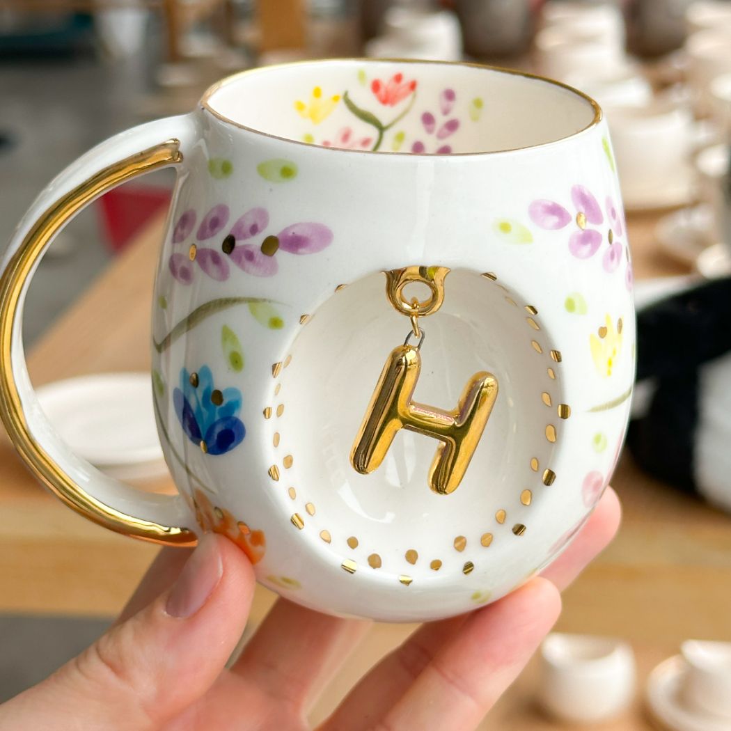 Porcelain mug with gold handle, floral pattern, and raised gold letter H on the front