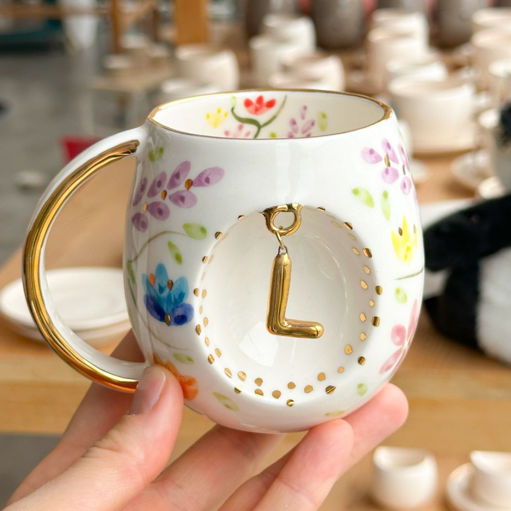 Porcelain mug with gold handle, floral pattern, and raised gold letter L on the front