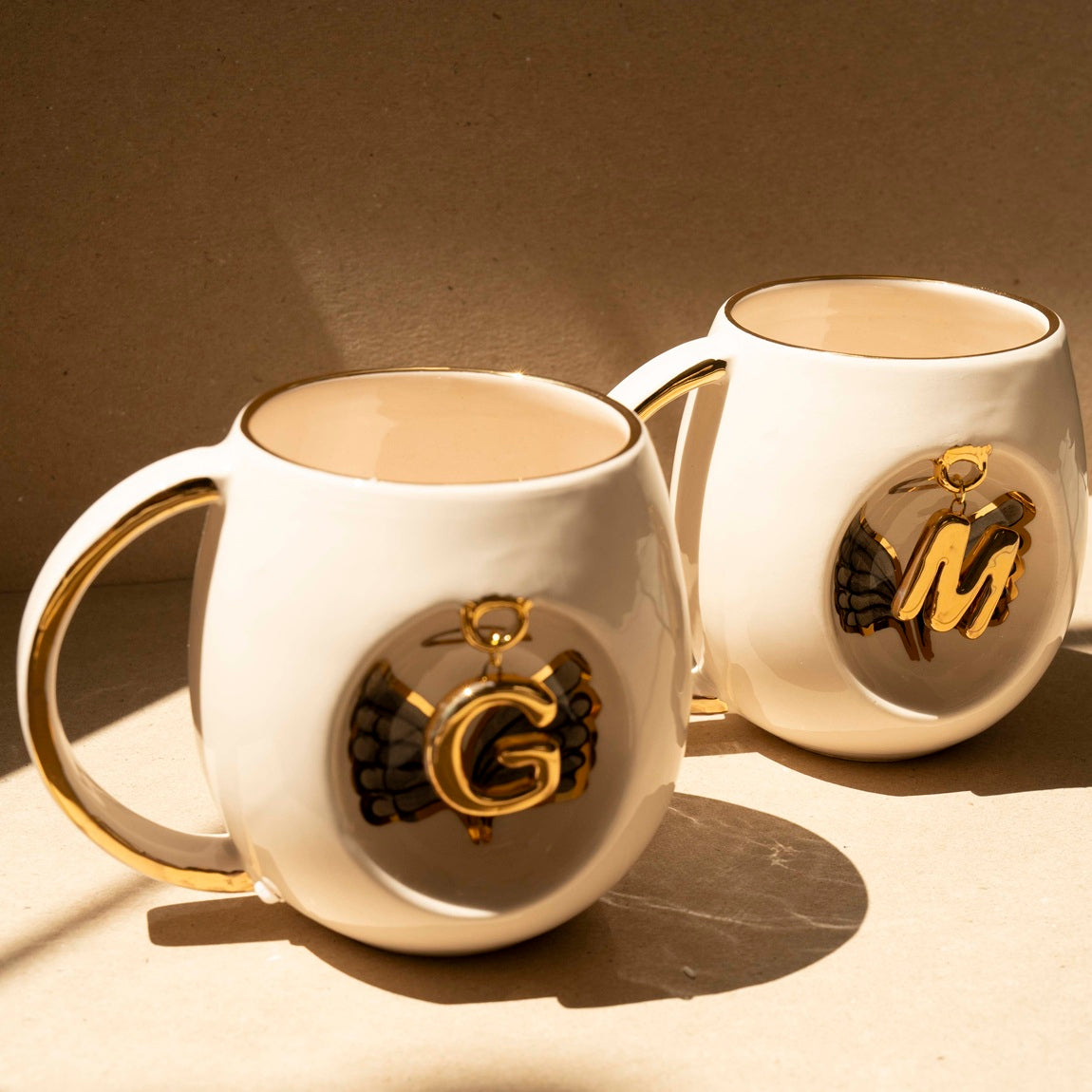 Pair of white porcelain mugs with gold handles and gold letter charms with black wings