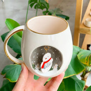White ceramic mug with gold handle and rim, snowman holding scarf on snowy scene