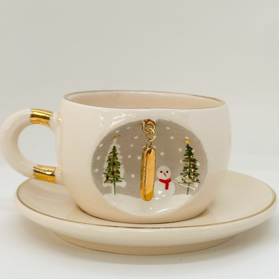Porcelain tea cup and saucer set, white with gold letter charm, snowman, and trees