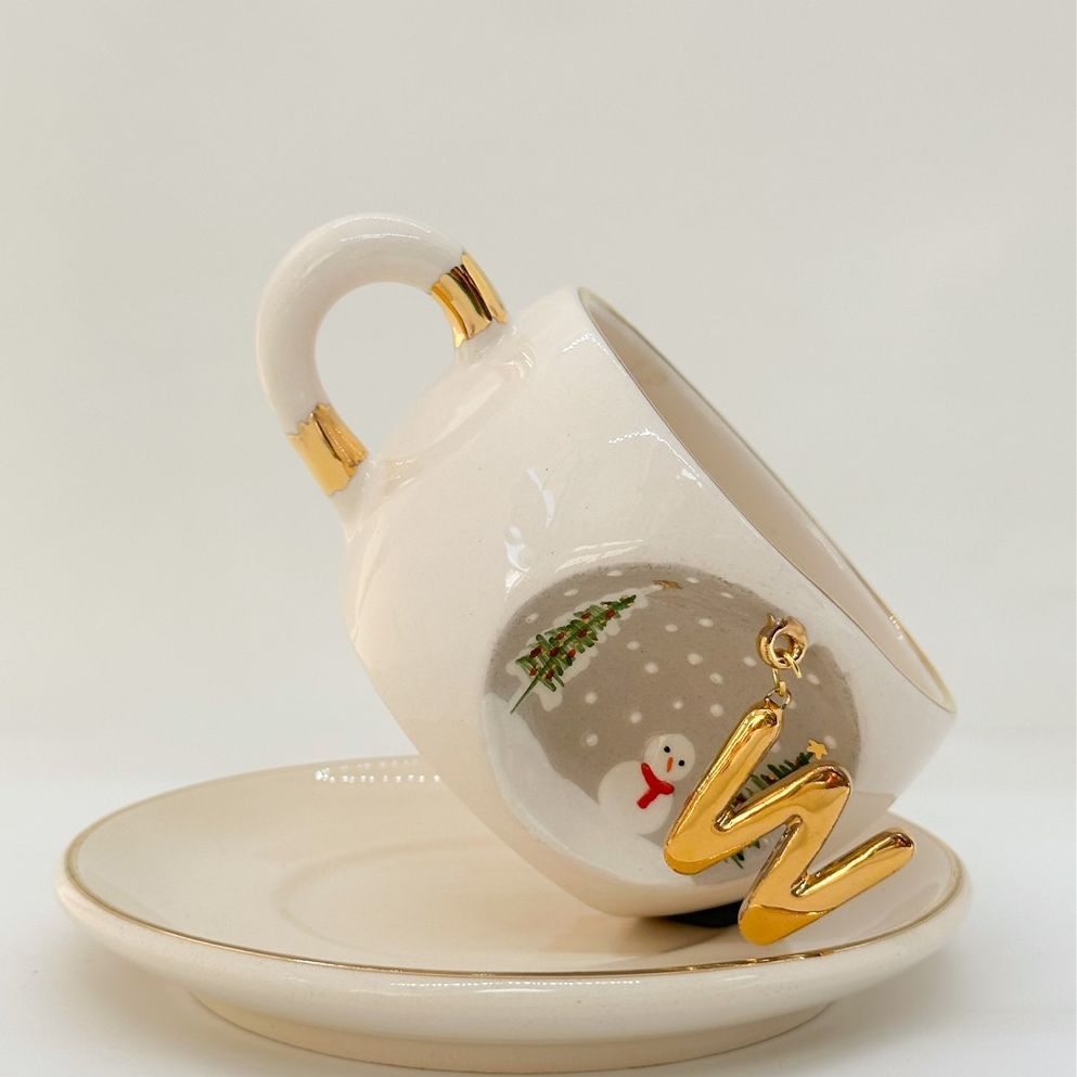 White porcelain tea cup on saucer, gold letter ornament with trees and snowman art, 160 ml.