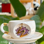 White ceramic mug with purple lotus and gold letter charm on saucer