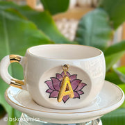 White ceramic mug with gold handle, hanging gold letter charm over pink lotus, on saucer