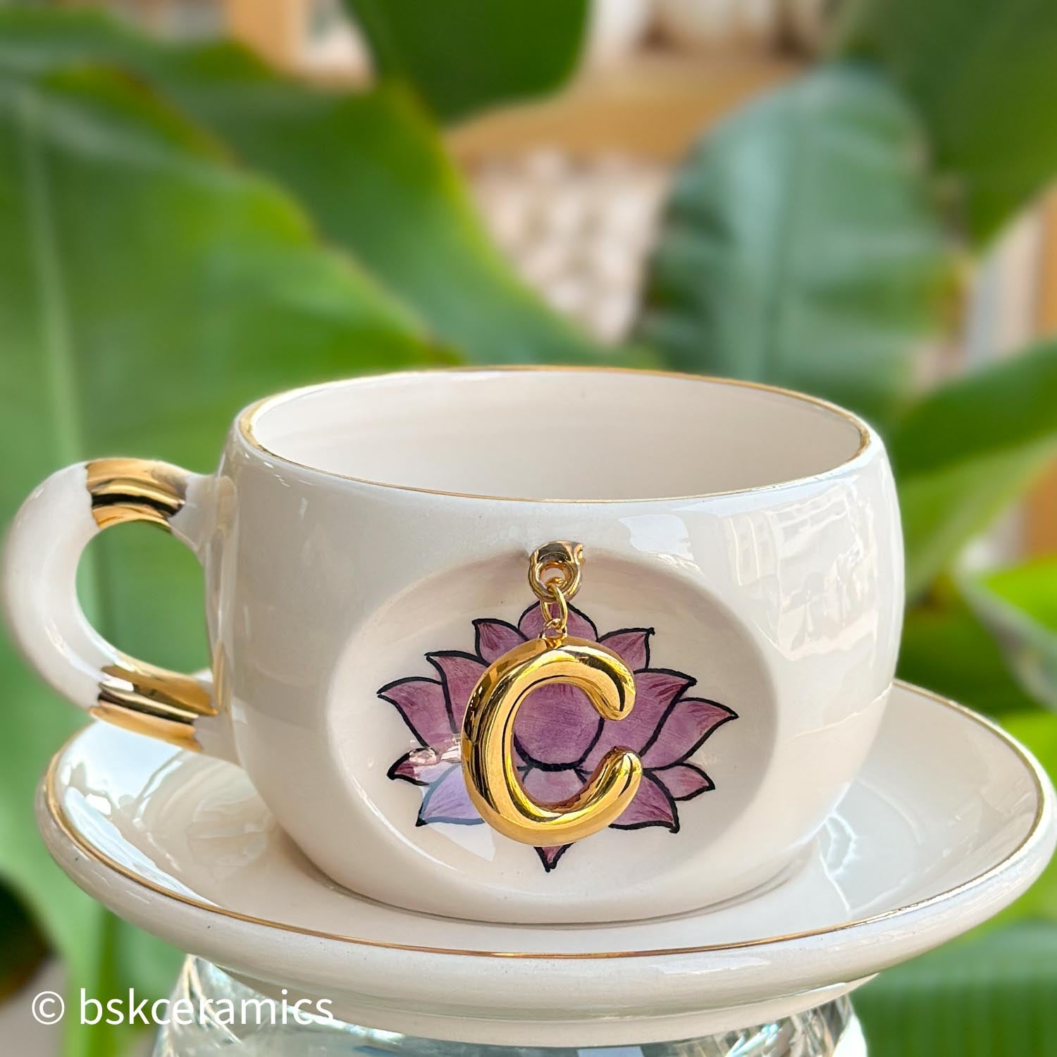White ceramic mug with gold handle and hanging gold letter charm over pink lotus design, on saucer