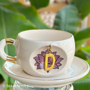 White ceramic mug with gold handle and hanging gold letter charm on pink lotus design, on saucer
