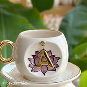 Ceramic coffee cup and saucer with gold handle, lotus letter A emblem on front