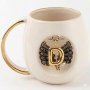 White porcelain mug with gold handle trim, black angel wings, and gold letter D pendant emblem.