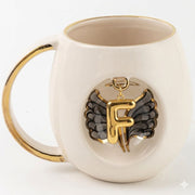 White porcelain mug with gold handle, black angel wings, and gold letter F pendant emblem.