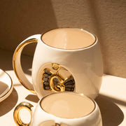 White porcelain mug with gold handle trim, black angel wings, and gold letter emblem.