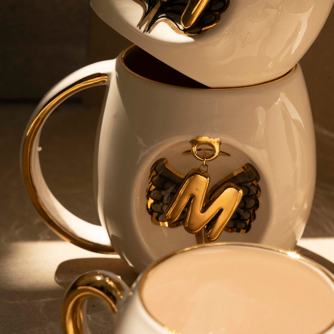 White porcelain mug with gold handle and gold letter charm featuring black wing details