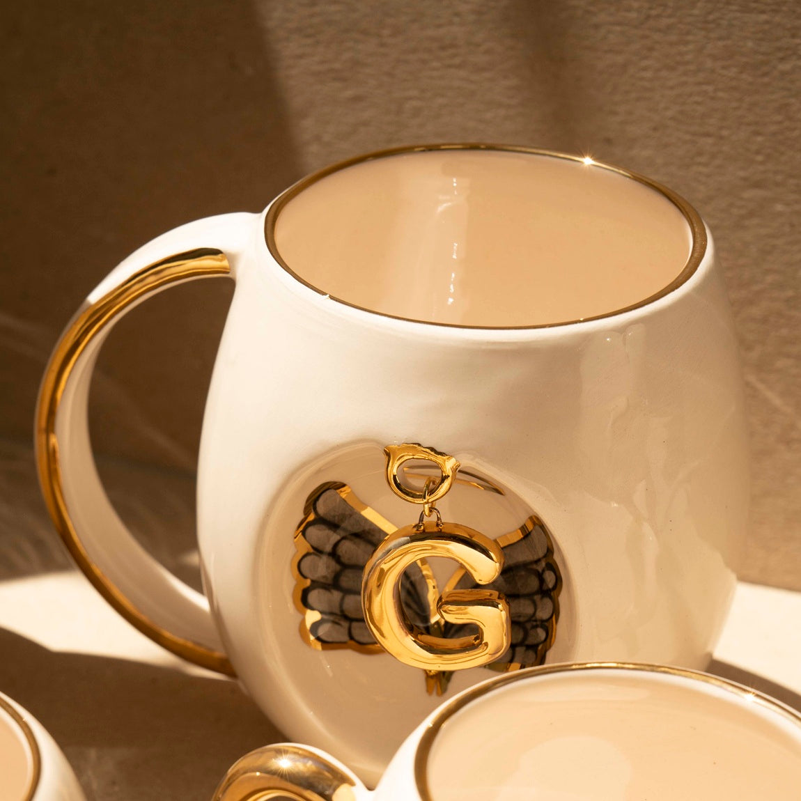 White porcelain mug with gold handle, gold letter charm, and black wing motif on front