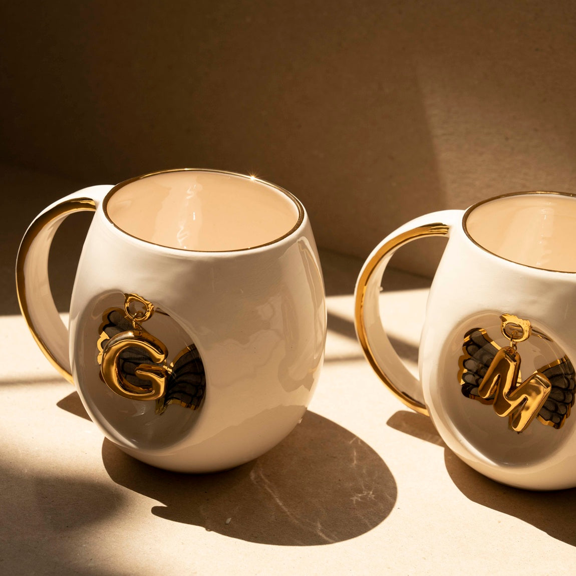 Two white porcelain mugs with gold handles and gold letter charms over black wing details