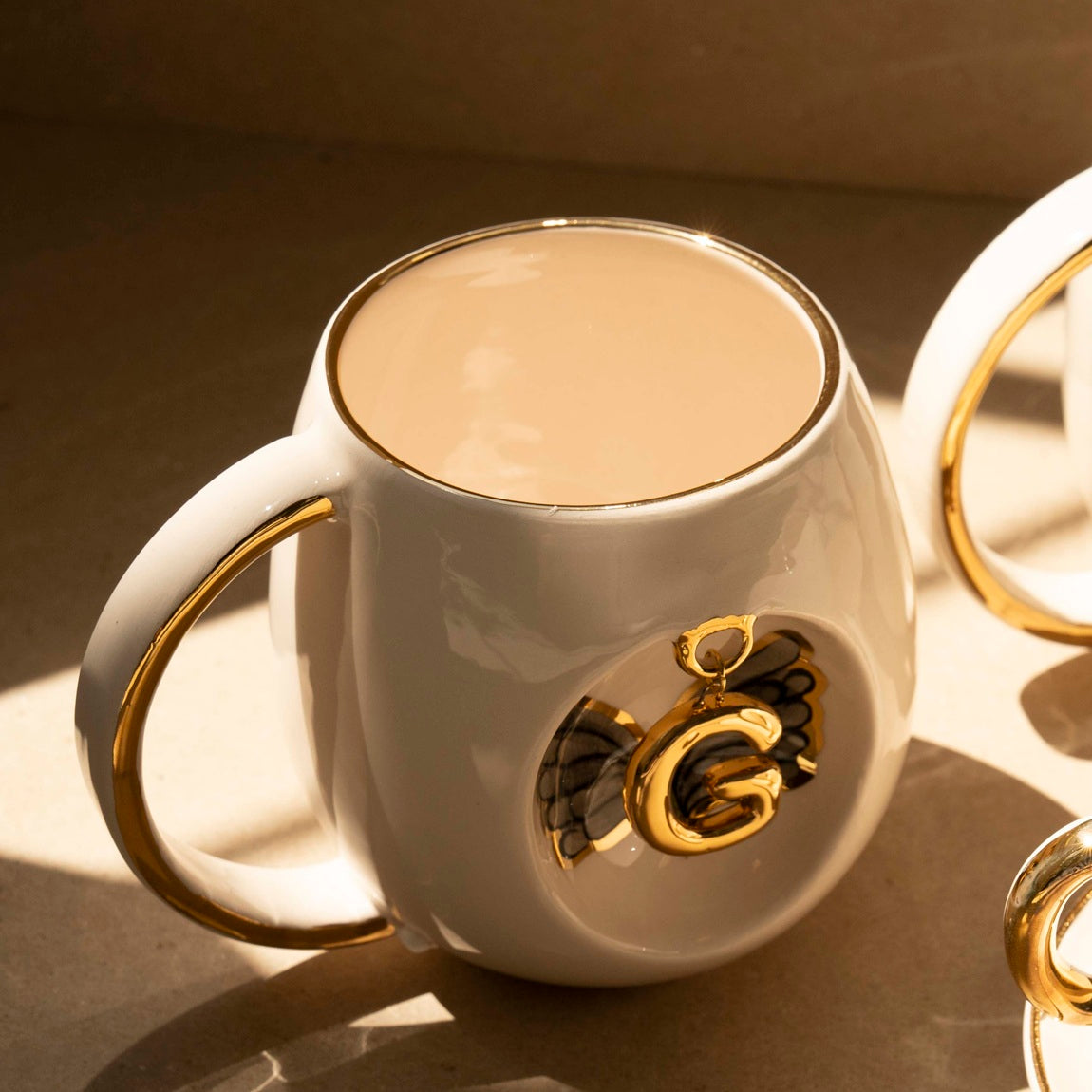 White porcelain mug with gold rim, black angel wing details, and gold letter emblem on front.