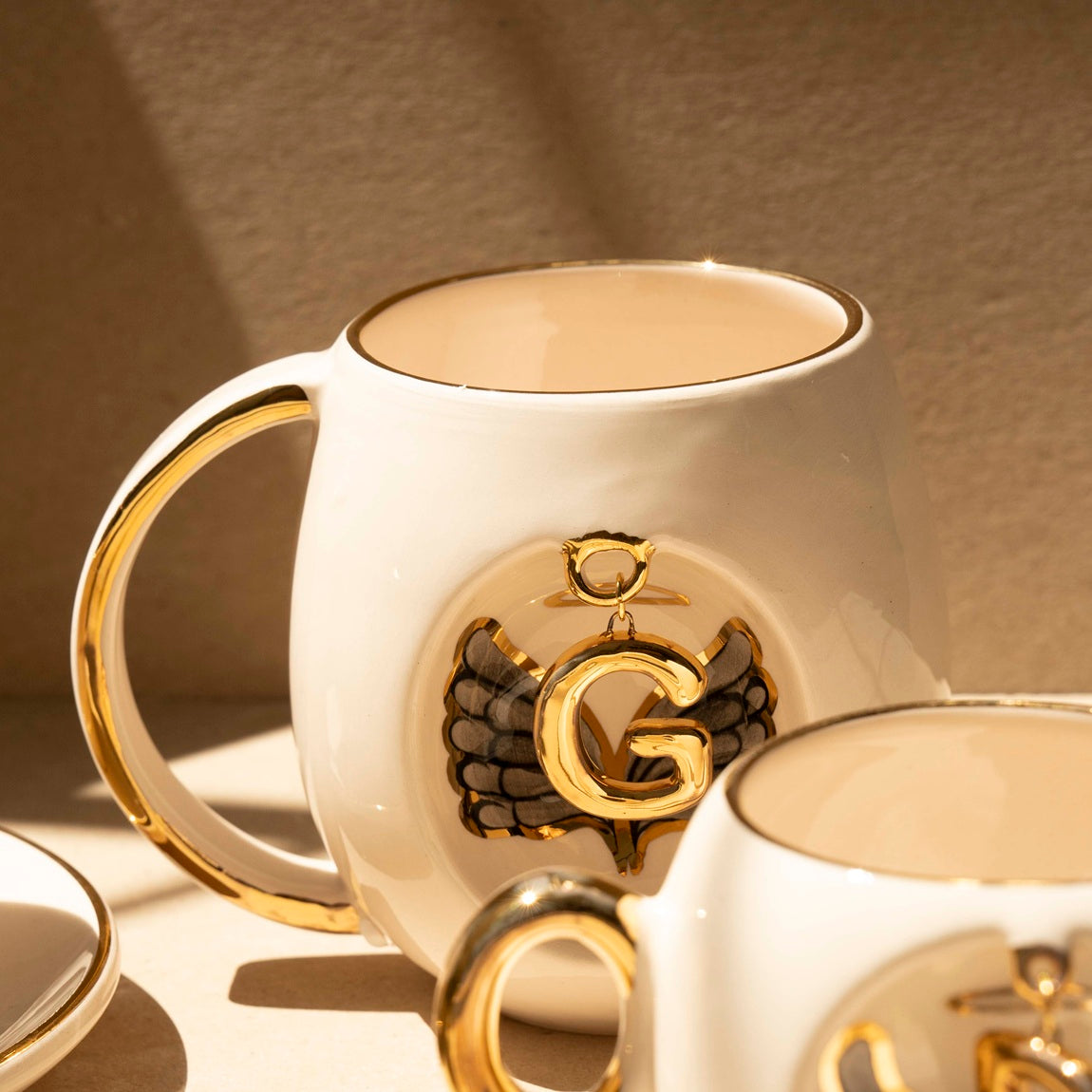 White porcelain mug with gold handle and gold letter charm with black wing artwork on front