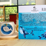 Ceramic coffee cup and saucer with blue whale illustration, gold handle; gift card included.
