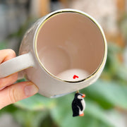 White ceramic mug with gold rim, empty interior, small penguin charm on side
