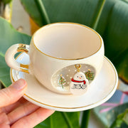 White tea cup with gold handle ring, snowman design and charm, shown above saucer