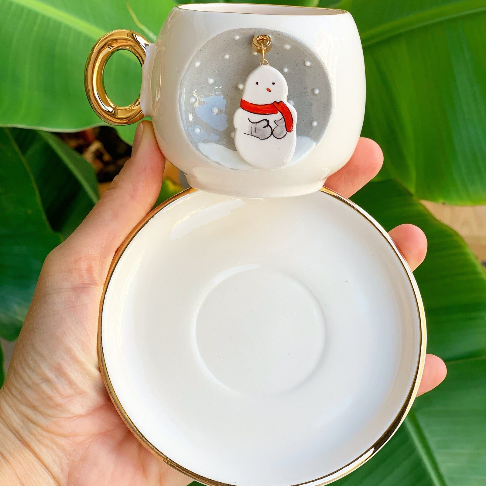 White ceramic snowman coffee cup with gold handle, gold rim, saucer, winter badge.