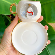 White ceramic snowman coffee cup with gold handle, gold rim, saucer, winter badge.