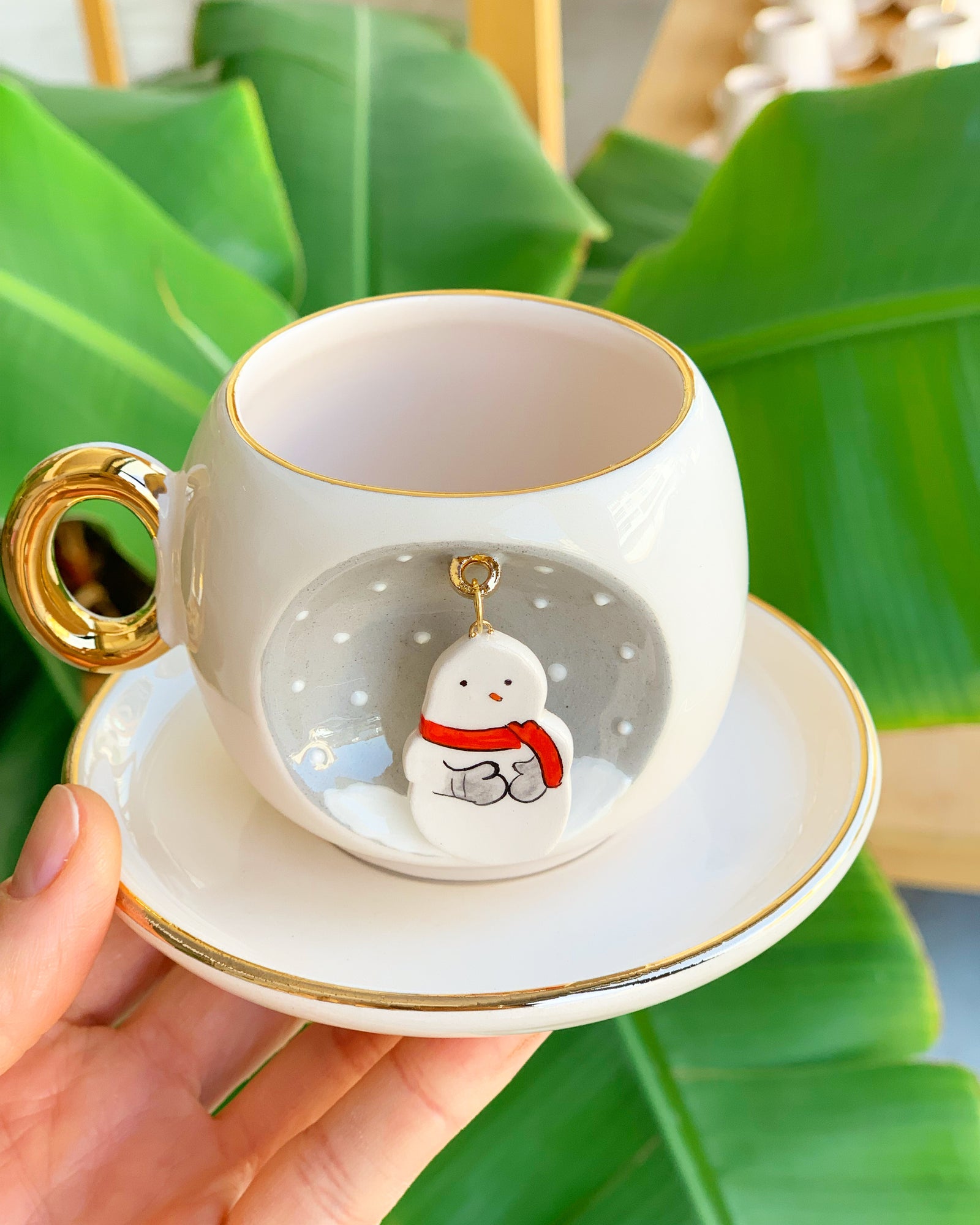 White ceramic coffee cup and saucer with gold rim, gold handle, snowman ornament on front.