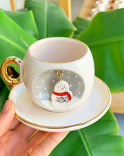 White ceramic coffee cup and saucer with gold rim, gold handle, snowman ornament on front.