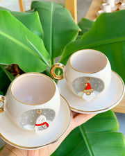 Pair of white ceramic snowman coffee cups on saucers with gold rims and handles, winter badges.