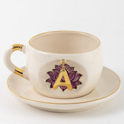 White ceramic mug on saucer with purple lotus and gold letter A charm