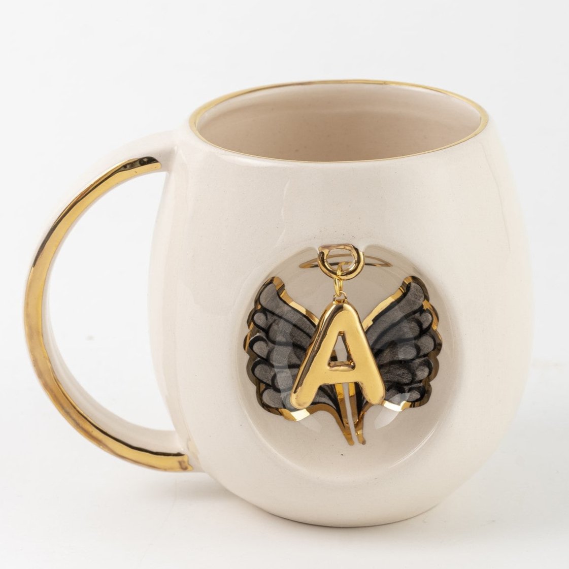 White porcelain mug with gold handle, black angel wings, and gold letter A pendant emblem.