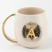 White porcelain mug with gold handle, black angel wings, and gold letter A pendant emblem.