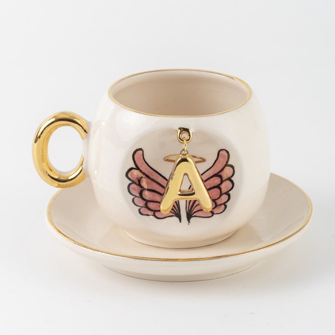 Ceramic coffee cup with gold handle, pink winged letter A charm and saucer.