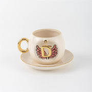 Ceramic coffee cup with gold handle, pink winged letter D charm and saucer.