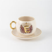 Ceramic coffee cup with gold handle, pink winged letter E charm and saucer.