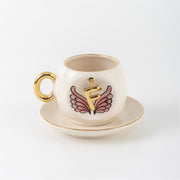 Ceramic coffee cup with gold handle, pink winged letter F charm and saucer.