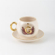 Ceramic coffee cup with gold handle, pink winged letter G charm and saucer.