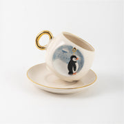 Ceramic coffee cup with penguin charm and blue splash print, gold handle, on saucer