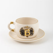 Porcelain mug and saucer with gold-toned angel wing letter emblem on front