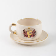 White mug with gold-rimmed opening and saucer, showing angel-wings letter print